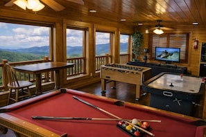 A pool table, foosball table, and television in a room with wooden walls and large windows.
