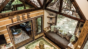 Un chalet chaleureux doté d’une grande fenêtre, d’une enseigne en bois sur laquelle est écrit le mot « ARBRE », d’un coin salon confortable avec des coussins et d’une décoration intérieure rustique.