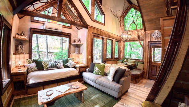 Un intérieur de chalet chaleureux avec un lit, un canapé et un coin repas, agrémenté de poutres en bois, de grandes fenêtres et d’une esthétique rustique.