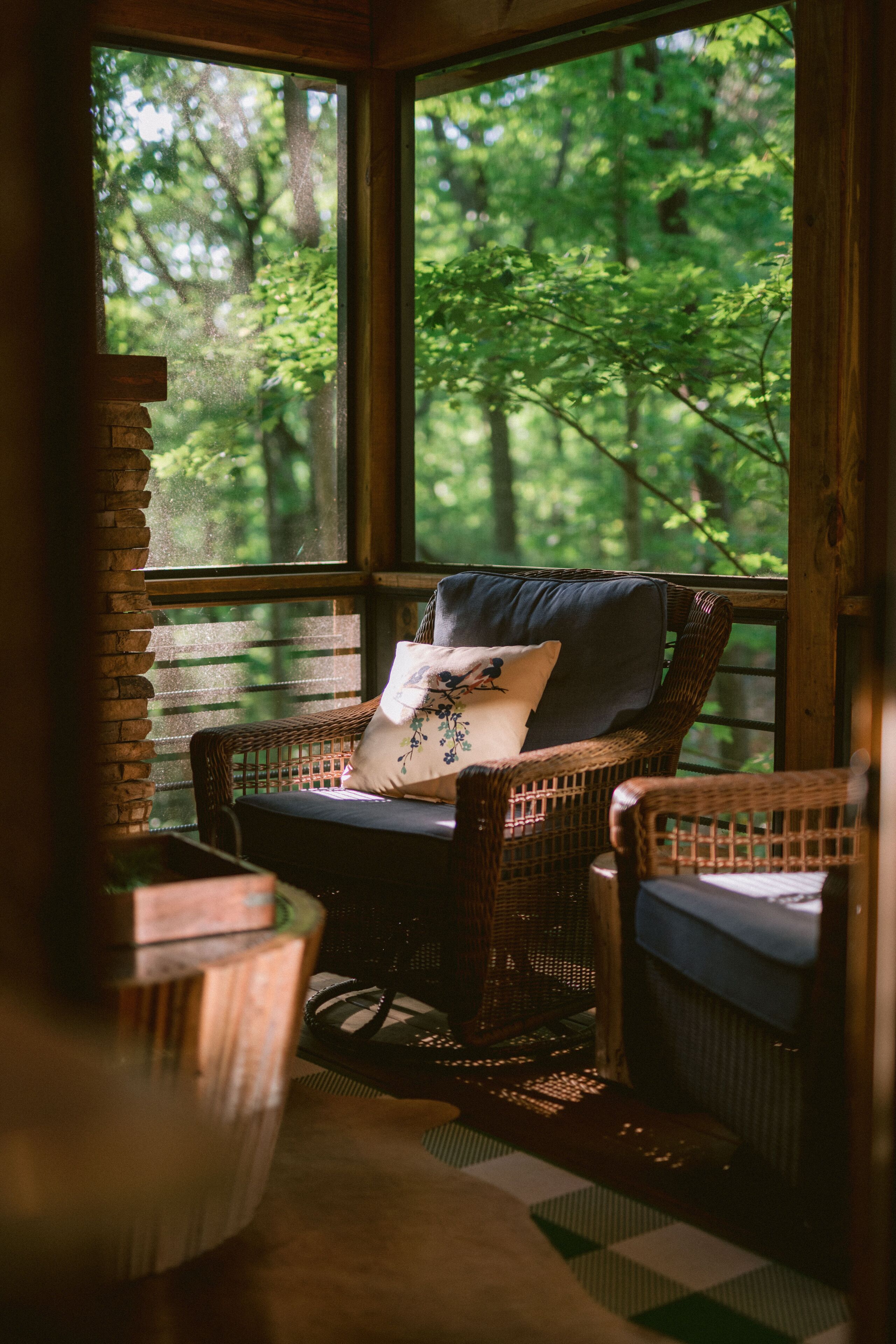A cozy wooden cabin with a wicker chair by a large window overlooking a forest.
