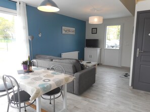 A modern living room with a grey sofa, a dining table with chairs, a TV on a cabinet, and a blue accent wall. - Gite NEW COMFORT-classified 3 *** for 2/4Pers, quiet. MER Prox. ANCV / WIFI (pluduno)