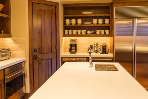 A kitchen with a wooden door, a coffee machine, a stainless steel refrigerator, and a built-in oven.