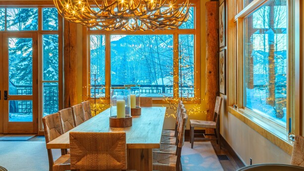 A dining area with a wooden table and chairs, large windows with a view of snow-covered trees, and a unique chandelier. -  5 Bed/5.5 Bath Ski-in Ski-out Lodge on Granite Ridge (Jackson)