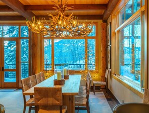 A dining area with a wooden table and chairs, large windows with a view of snow-covered trees, and a unique chandelier. -  5 Bed/5.5 Bath Ski-in Ski-out Lodge on Granite Ridge (Jackson)