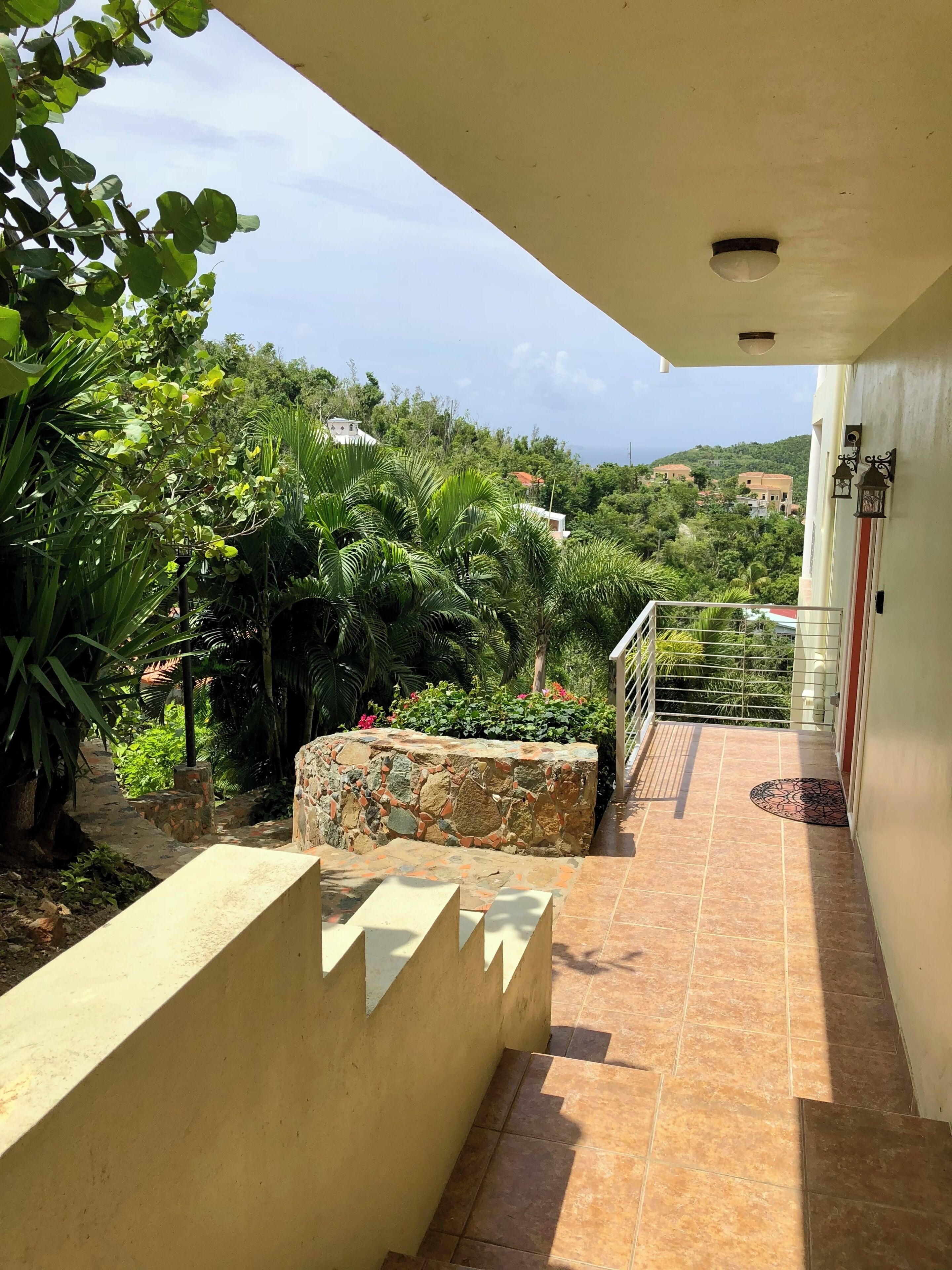 Un balcon doté d’un mur en pierre, d’une rambarde en métal et offrant une vue sur un jardin luxuriant et des bâtiments éloignés.