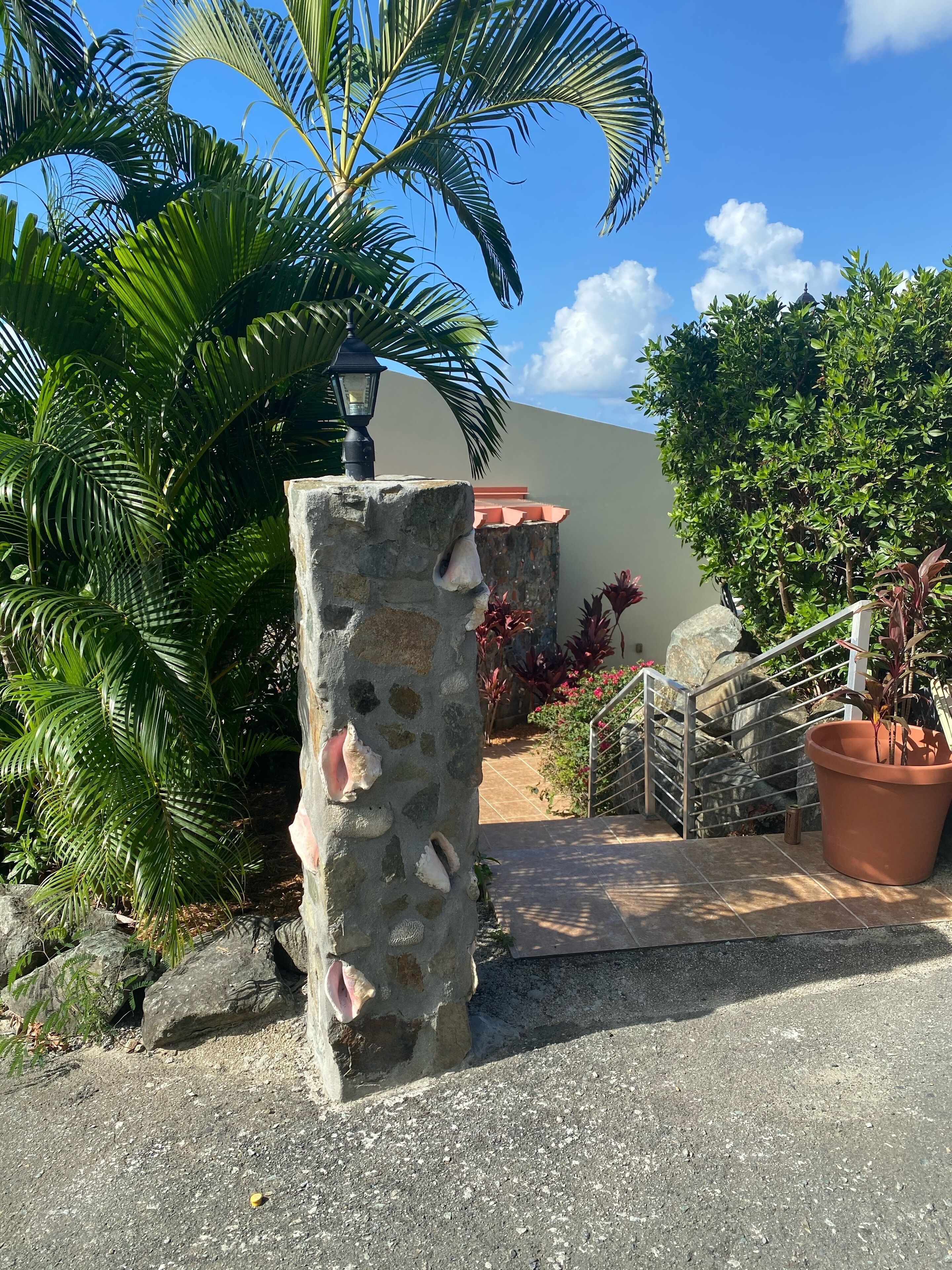 A stone pillar with a lamp, surrounded by tropical plants and a staircase leading to a building.