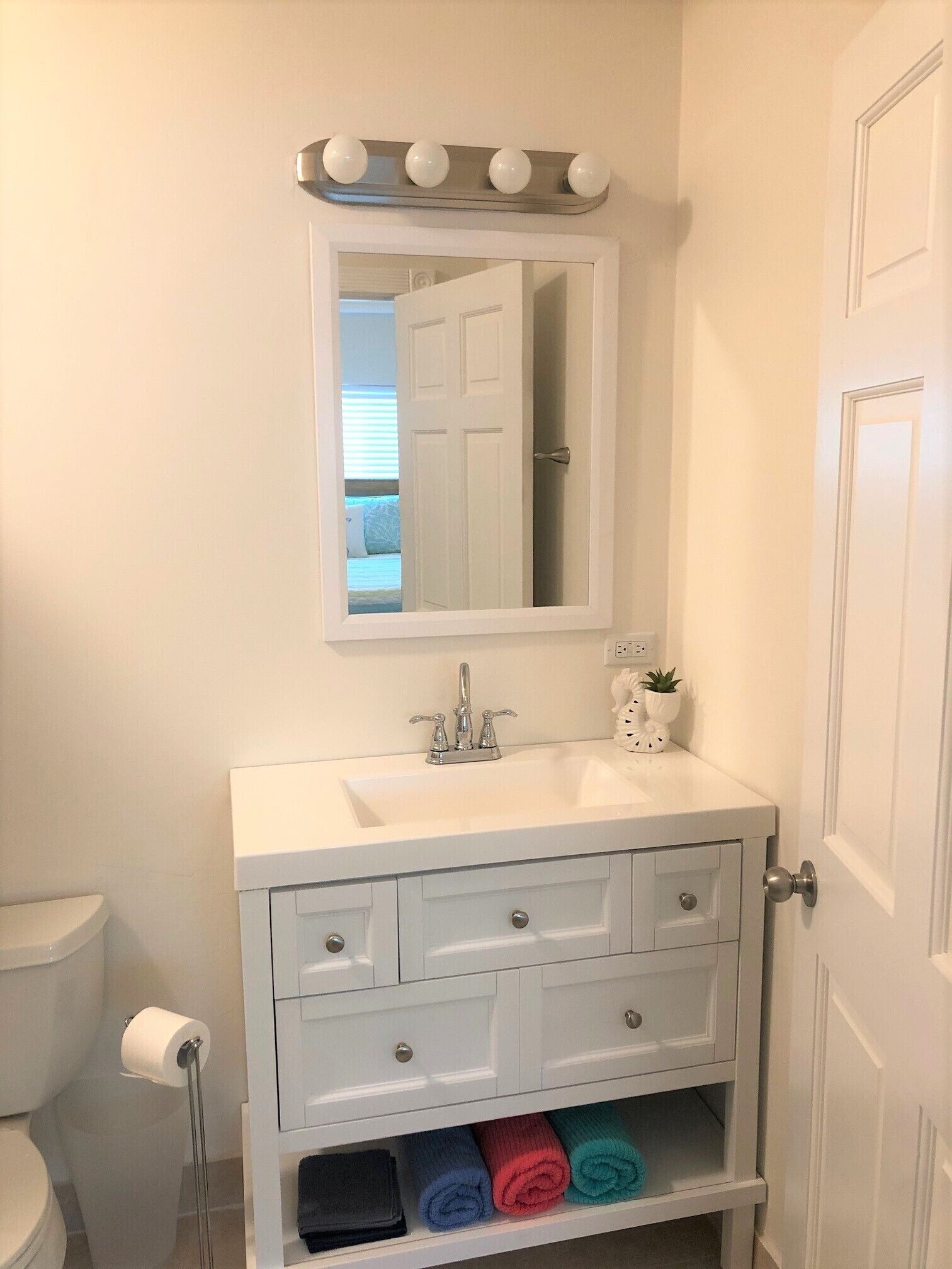 Une salle de bain avec un meuble-lavabo blanc, un miroir avec éclairage intégré, des toilettes et une étagère avec des serviettes.