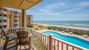 A balcony with a pool, beach view, and outdoor seating.