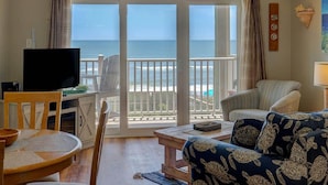 A living room with a television, a dining table, and a sofa. There is a view of the ocean through large sliding glass doors.