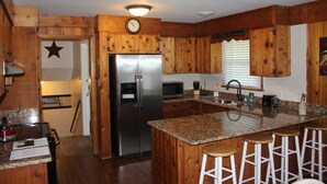 A kitchen with wooden cabinets, a stainless steel refrigerator, a granite countertop, and a bar area with stools.