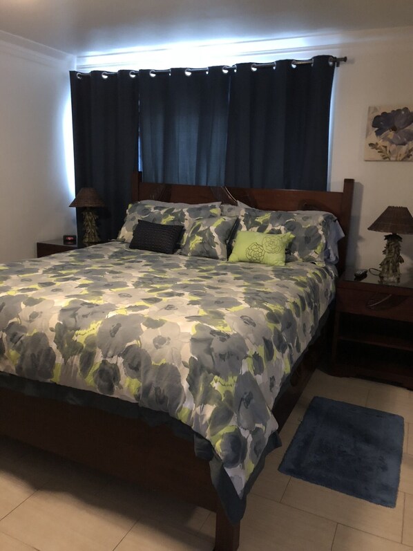 Een bed met een bloemenbedekking, een nachtkastje met een lamp en een blauw vloerkleed op een betegelde vloer.
