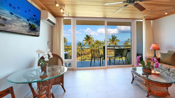 Een ruime kamer met een glazen eettafel, rieten stoelen, een bank en een balkon met uitzicht op palmbomen en de oceaan.