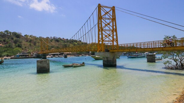 A bridge with a yellow truss structure spanning over a body of water with boats. - Ceningan Inn (Ceningan Island)
