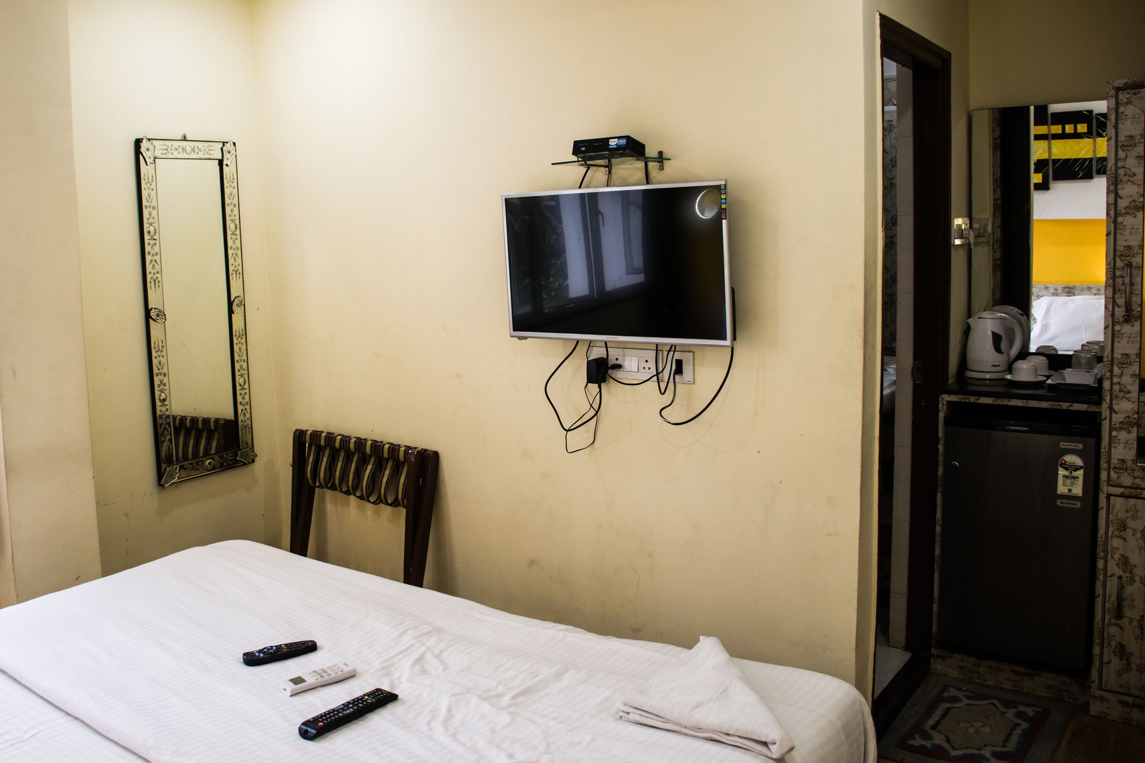 A hotel room with a bed, a television mounted on the wall, a mirror, a chair, and a small kitchenette area.