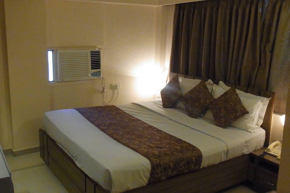 A hotel room with a bed, a bedside table, a lamp, and an air conditioning unit.