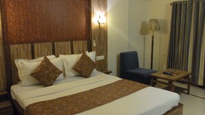 A hotel room with a large bed, two pillows, a bedside table, a chair, a lamp, and a curtain. - Hotel Eden (Ahmedabad)