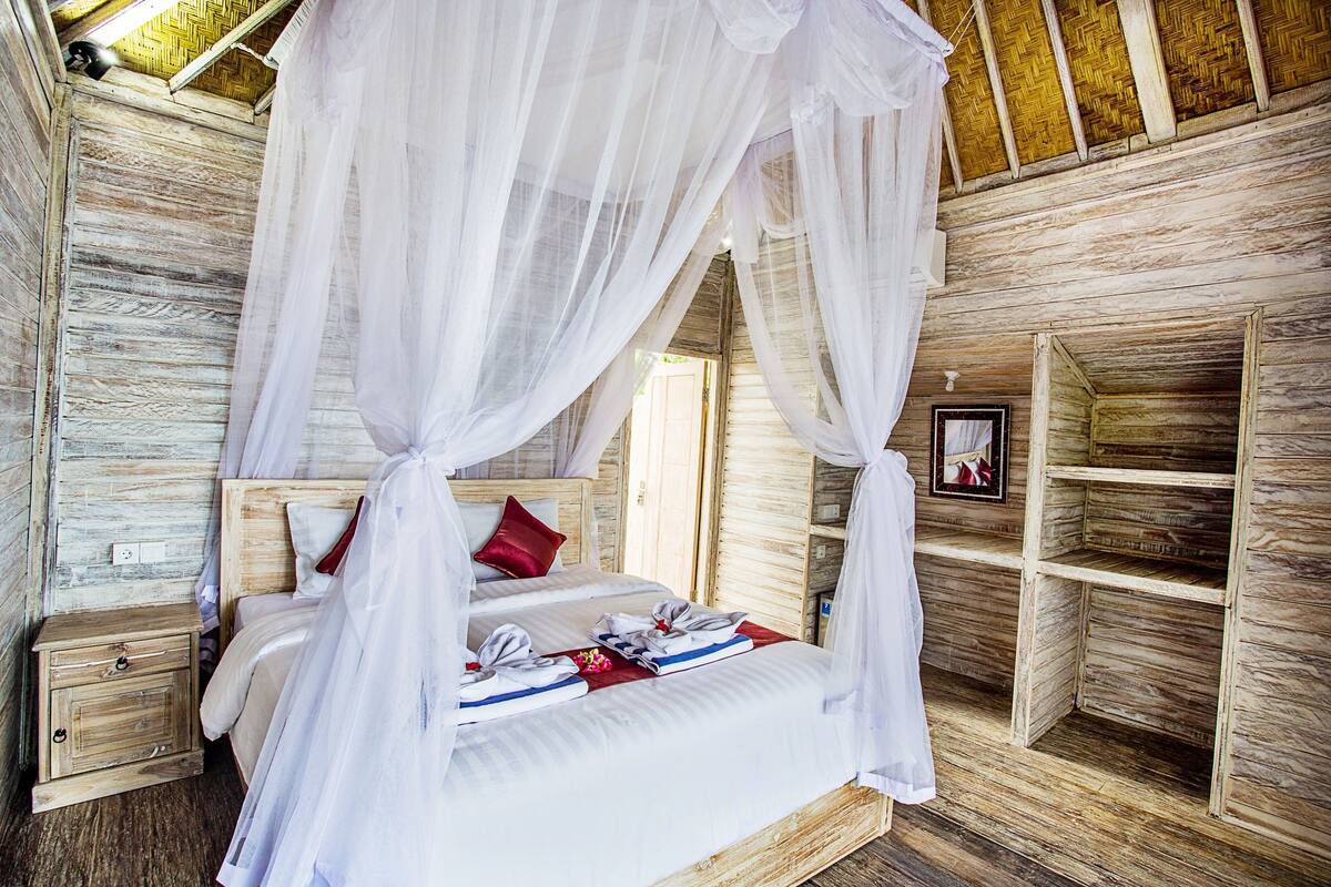 Een houten kamer met een bed onder een hemelbed, een nachtkastje en een plank met een ingelijste foto.