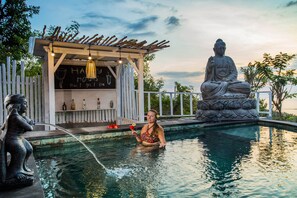 Uma área de piscina com uma estátua de Buda, uma estrutura de madeira com uma placa e uma pessoa na água.