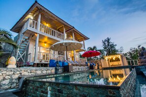 A two-story wooden building with a balcony, a swimming pool, and outdoor seating with umbrellas.