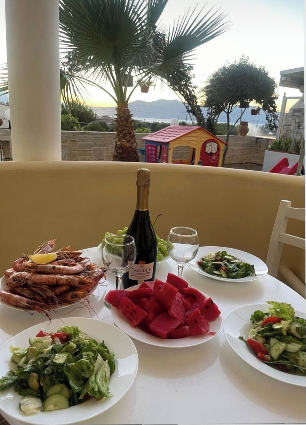 A table set with seafood, salads, and a bottle of wine, overlooking a scenic view with palm trees and a small building.