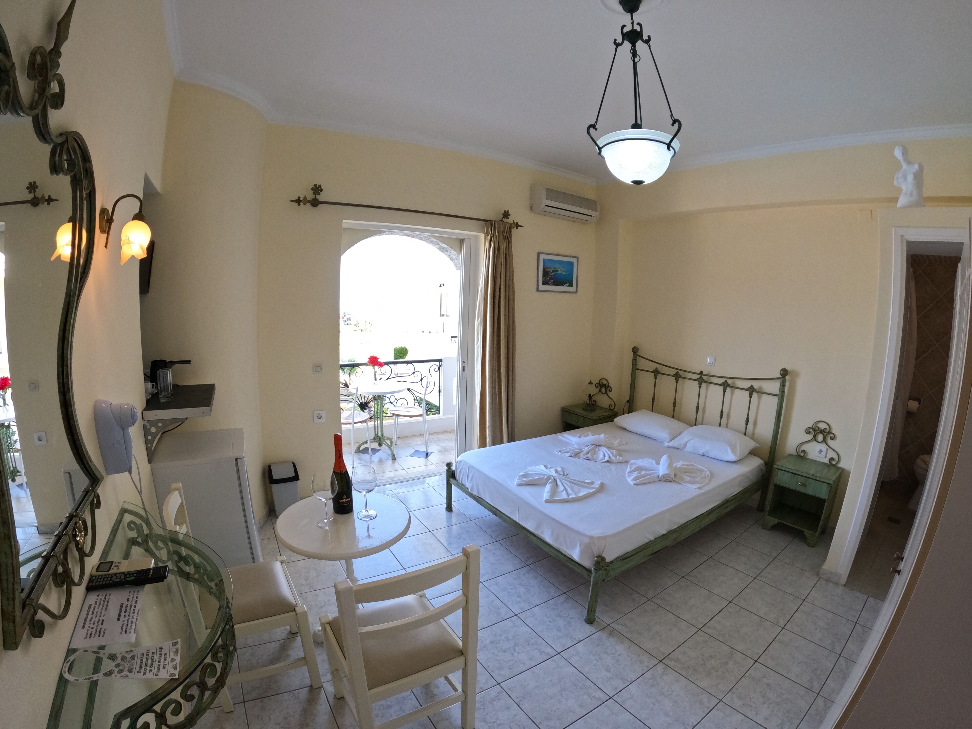 A bedroom with a bed, a small table with a bottle and glass, a chair, a mirror, and a balcony with a view.
