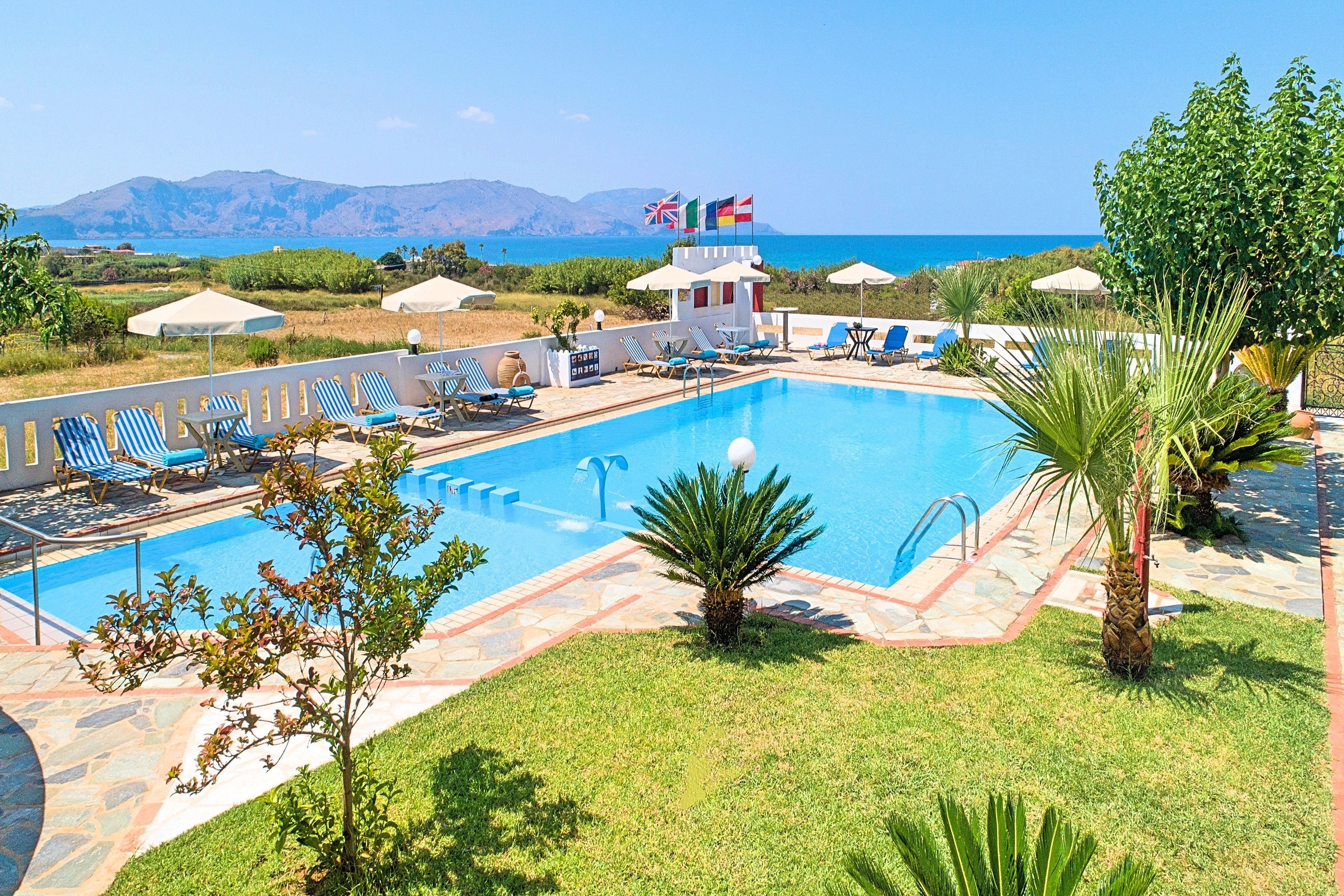 A swimming pool with lounge chairs and umbrellas, flags, and a view of the sea and mountains.