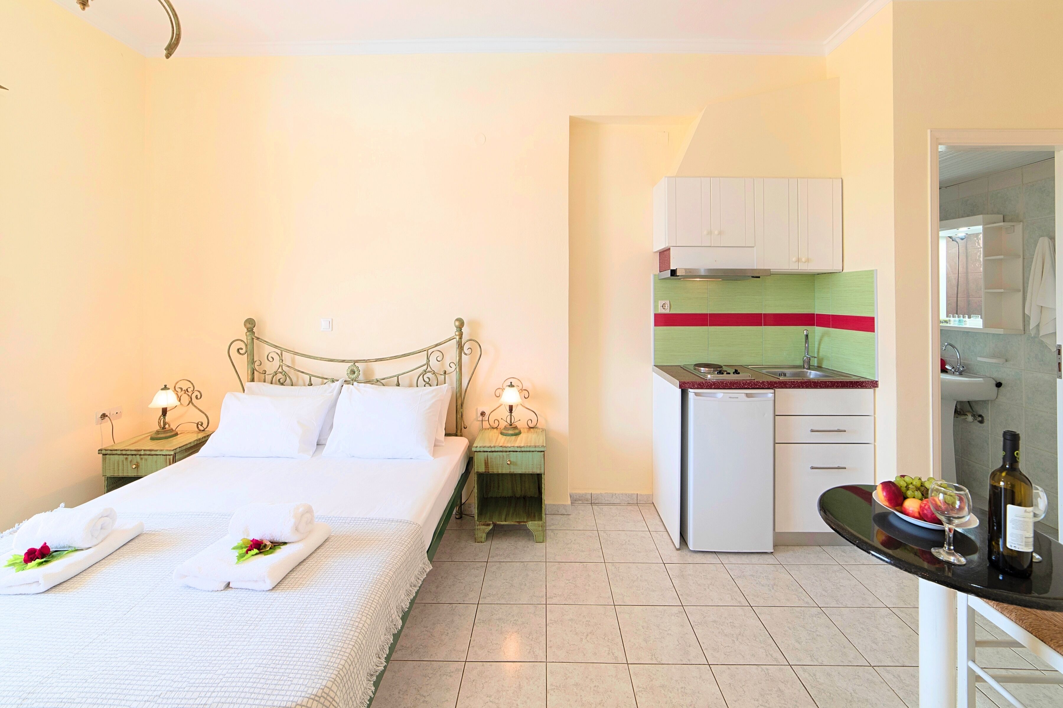 A hotel room with a bed, two bedside tables with lamps, a small kitchenette, and a bathroom visible through an open door.