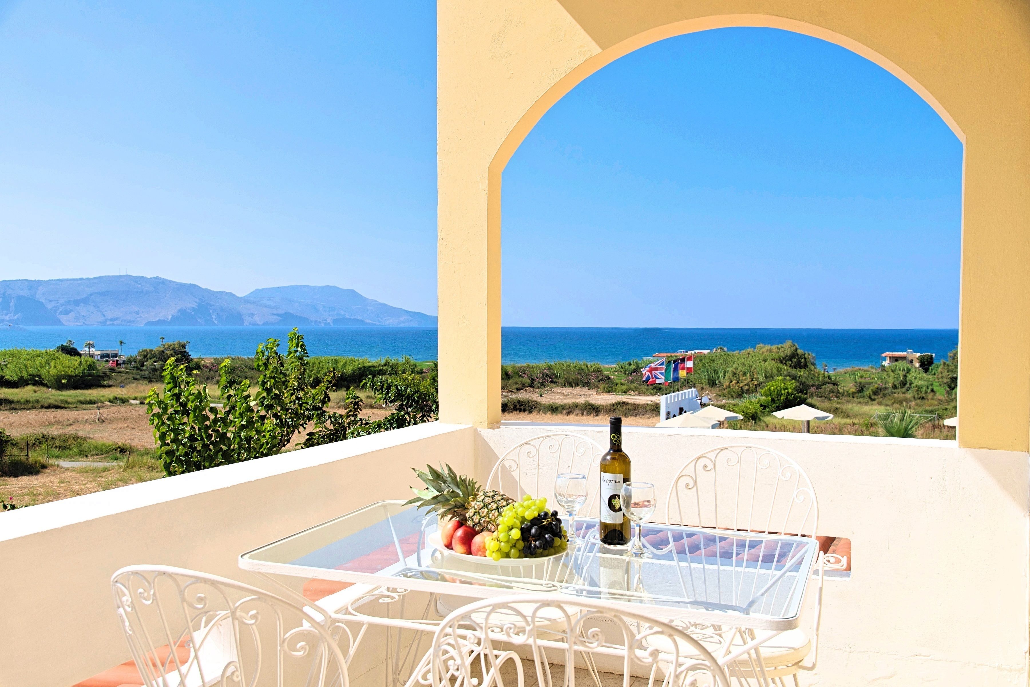 A balcony with a table set for two, a bottle of wine, and a bowl of fruit. The view includes a beach and distant mountains.