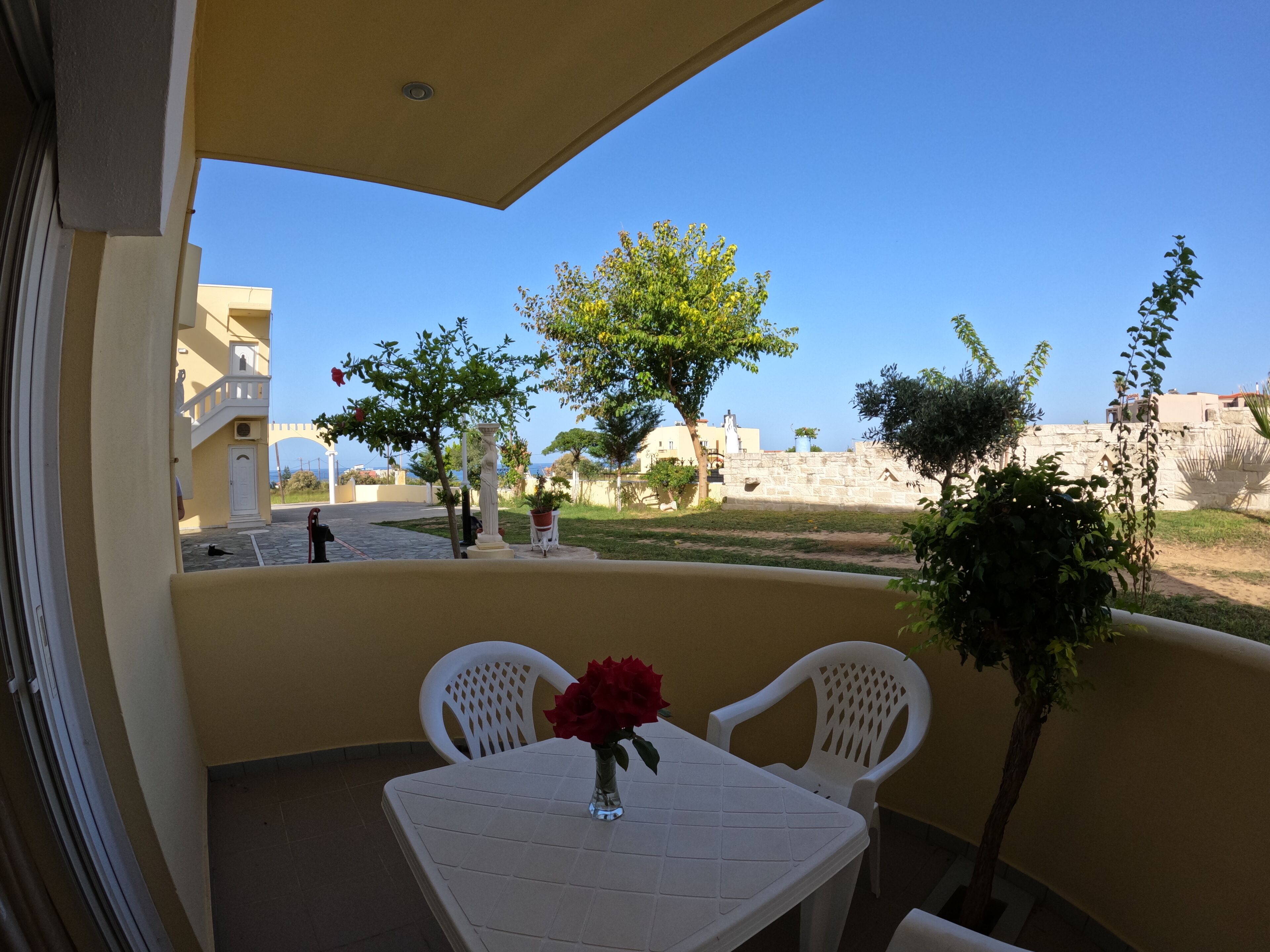 A balcony with a table and chairs, a vase with a red flower, and a view of the sea and buildings.