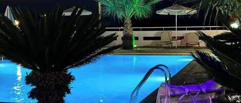 Zona de piscina nocturna con vista despejada al agua, una palmera y una silla iluminada con luz púrpura.