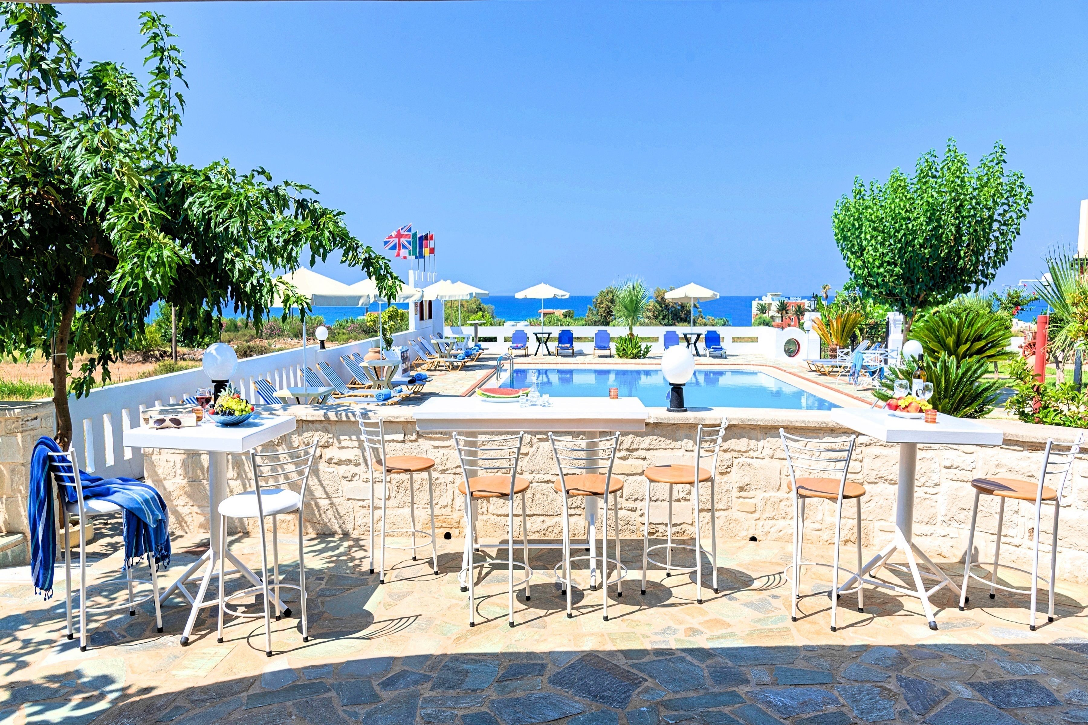 Uma área de bar à beira da piscina, com mesas e cadeiras brancas, piscina e vista desimpedida para o mar.