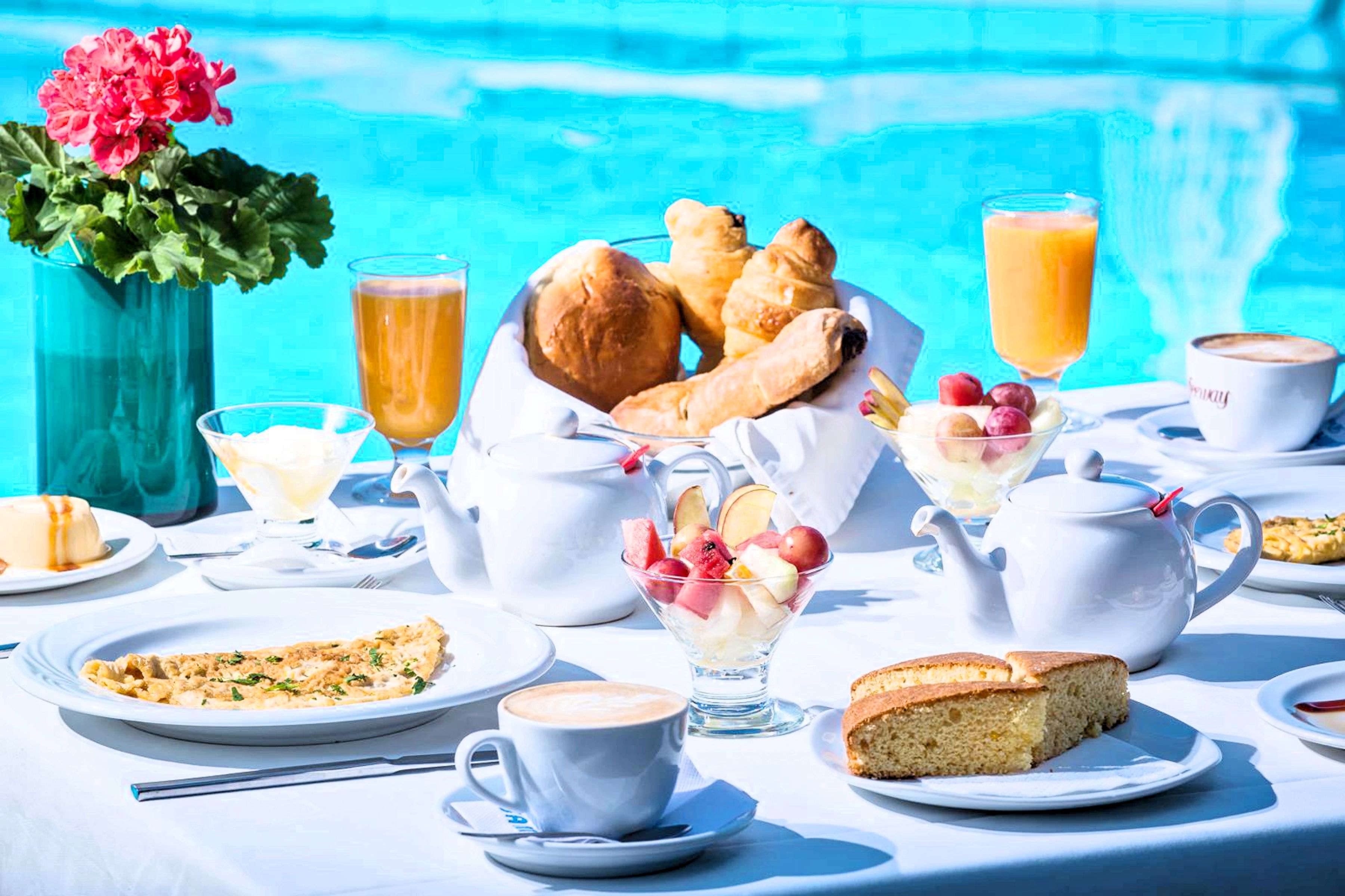 A table set with breakfast items including croissants, fruit, and coffee, near a pool.