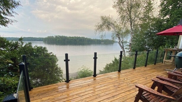 A wooden deck with a railing overlooking a lake, surrounded by trees. - Lake Joseph 3 Bedroom Cottage and Bunkie (MacTier)