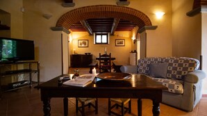 A living room with a TV, a table with a book and glasses, a patterned sofa, and a brick archway. - Villa with swimming pool, tennis court and table tennis ideal for sports lovers and the enjoyment of (Castiglion Fiorentino)