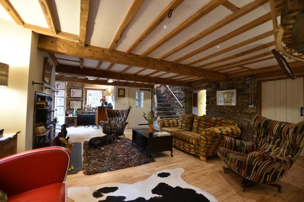 A rustic interior with exposed wooden beams, a stone wall, and a cozy seating area with patterned sofas and a red armchair.