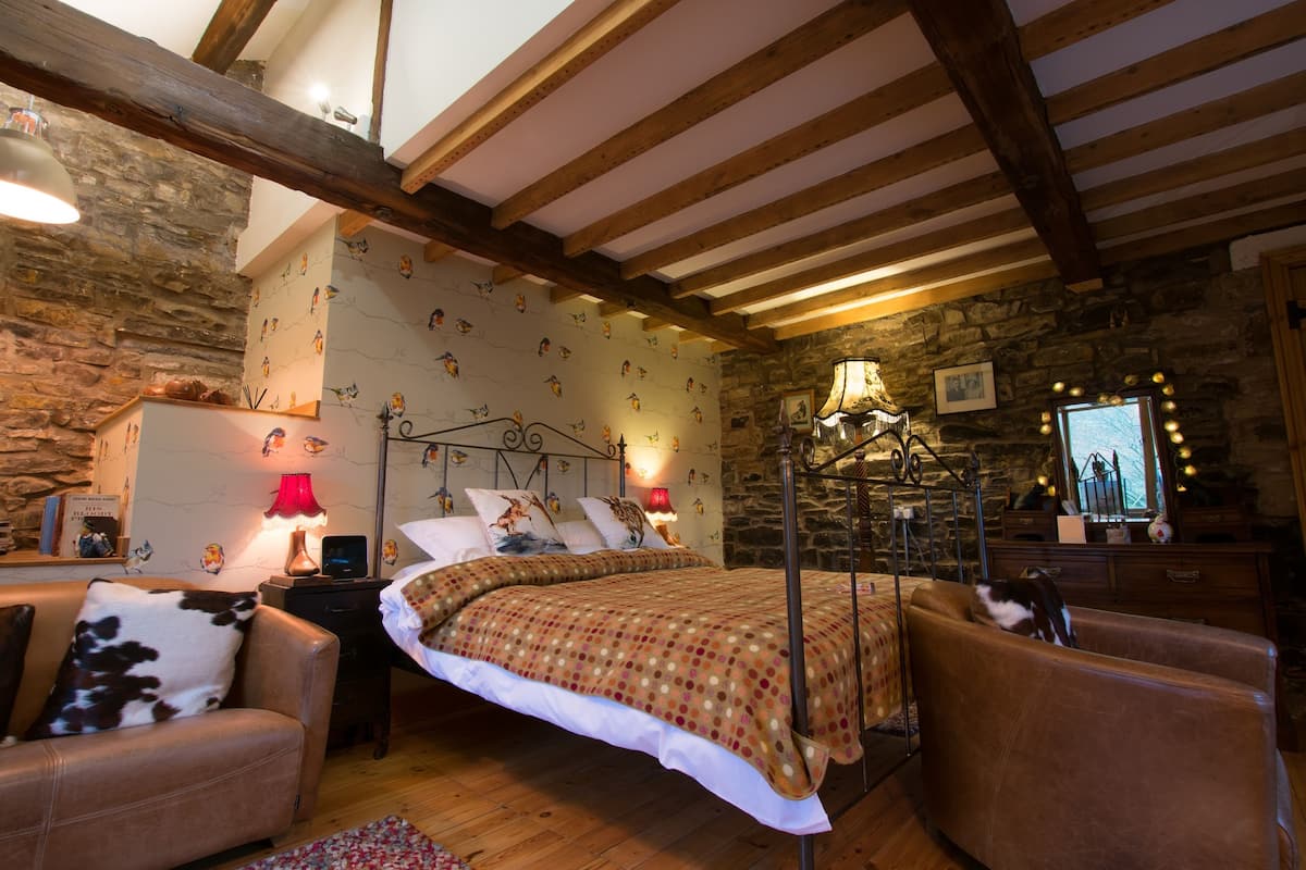 A cozy bedroom with a bed, a chair, a nightstand, a lamp, and a stone wall.