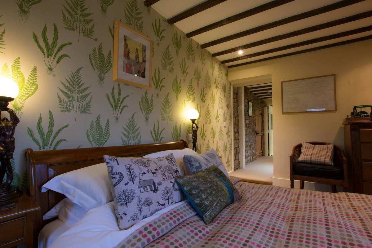 A bedroom with a bed, a nightstand, a lamp, a chair, and a wall with a fern pattern.