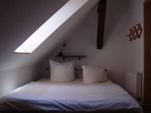 Una habitación pequeña, con poca luz y techo inclinado, cuenta con una cama individual con ropa blanca y una lámpara fijada a la pared.
