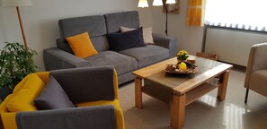 Ein Wohnzimmer mit grauem Sofa, gelben Sesseln, einem Couchtisch mit Früchten und einer Vase sowie einer Topfpflanze.