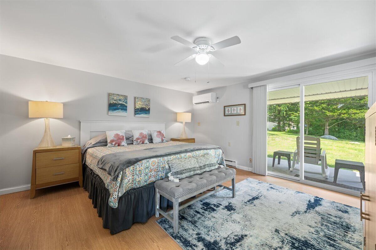 A bedroom with a bed, bedside lamps, a bench, a ceiling fan, and a view of the outdoors through a sliding glass door.