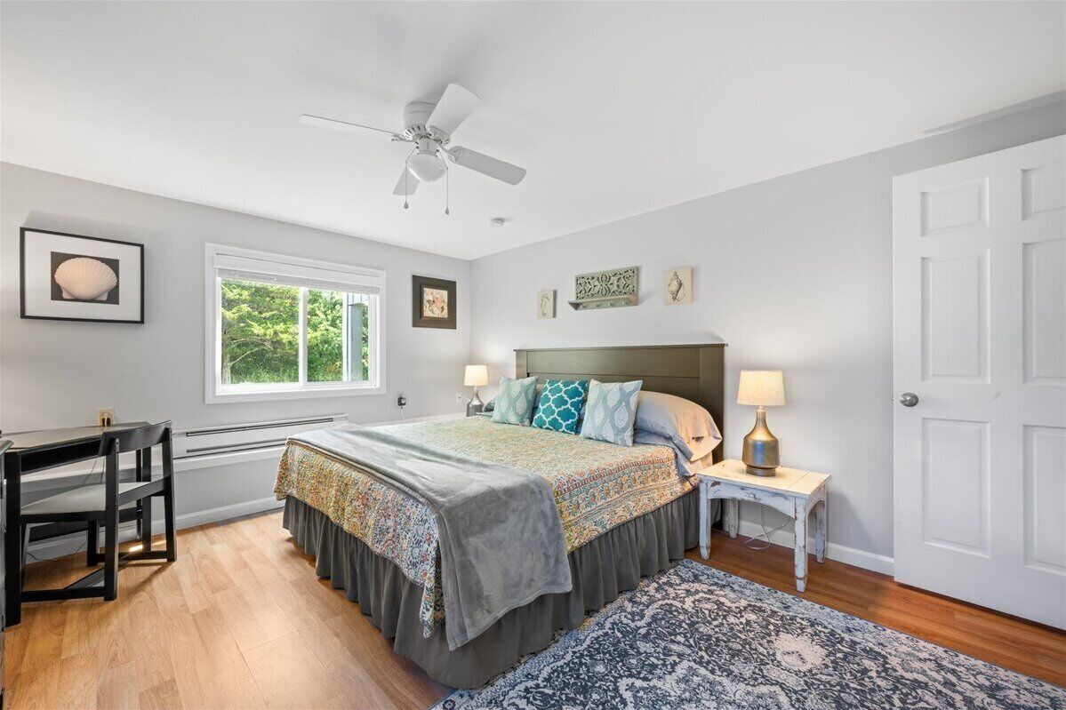 Une chambre à coucher comprenant un lit, un bureau, une chaise, une table de chevet, une lampe, un ventilateur de plafond et une porte.