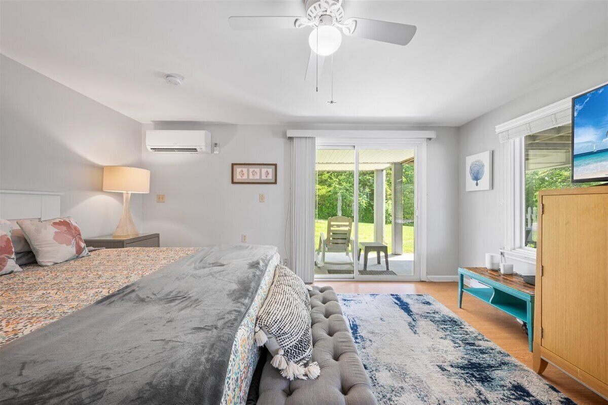 A bedroom with a bed, a ceiling fan, an air conditioner, a window with a view of greenery, and a wooden cabinet.