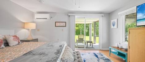 A bedroom with a bed, a ceiling fan, an air conditioner, a window with a view of greenery, and a wooden cabinet.