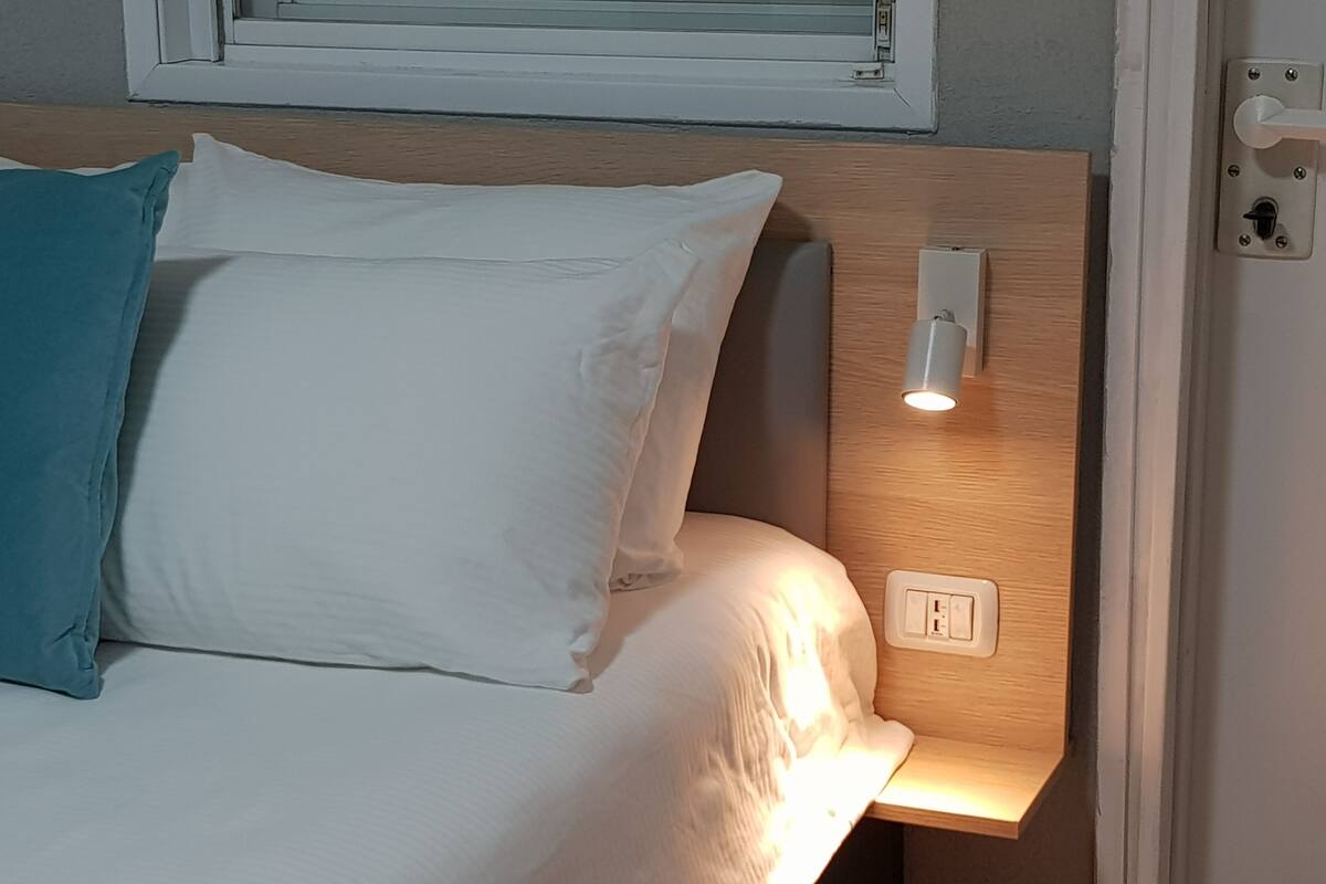 A bed with white bedding and a teal pillow, a bedside lamp, and a power outlet on the wall.