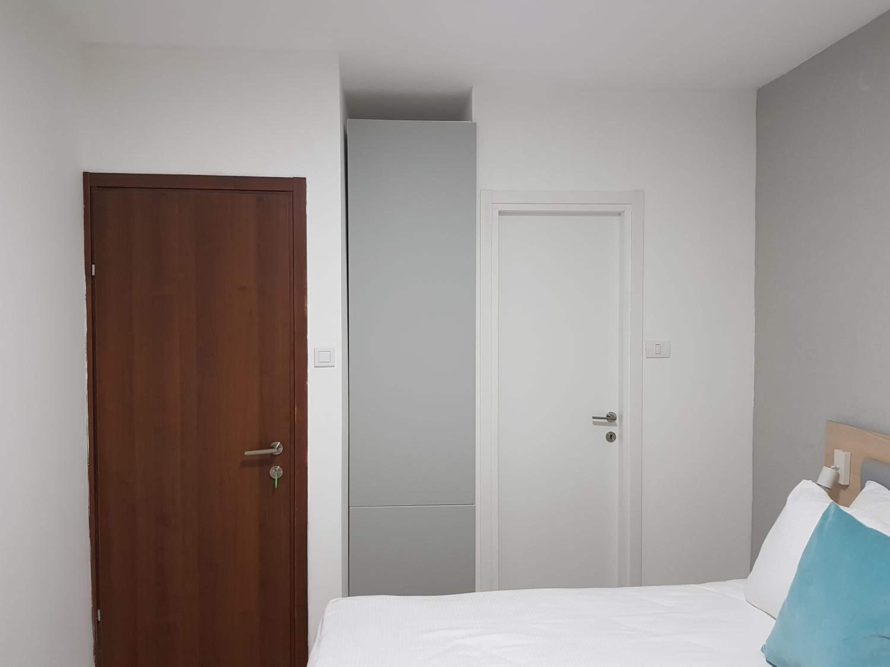 Quarto com porta de madeira, porta branca e cama com roupa de cama branca e almofada azul.