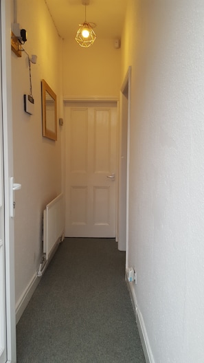 A narrow hallway with a white door at the end, a radiator on the left, and a wall-mounted light fixture. - Stunning Somerset House (Middlesbrough)