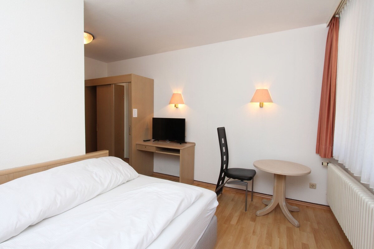 Ein Hotelzimmer mit einem Bett, einem Schreibtisch mit Fernseher, einem Sessel, einem kleinen Tisch und zwei Wandleuchten.