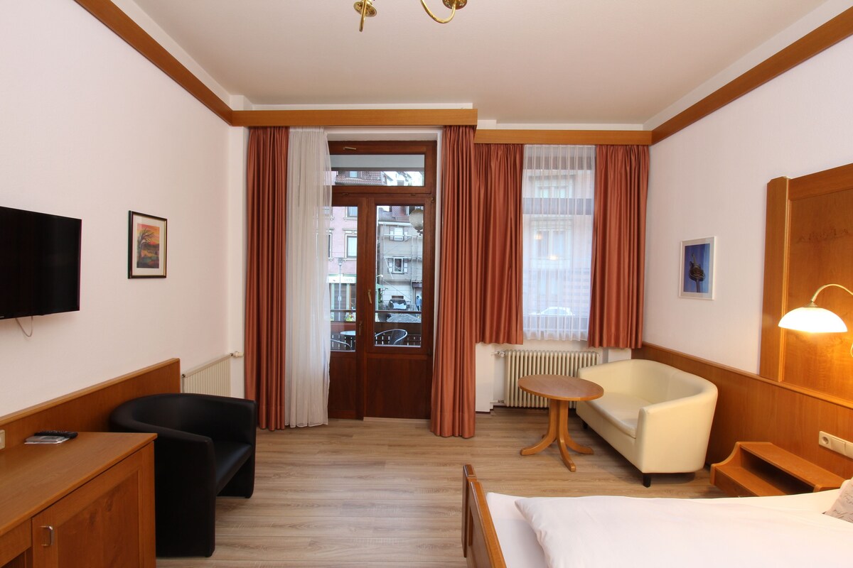 Ein Hotelzimmer mit Bett, Schreibtisch, Stuhl, Sofa, Fernseher und einem Fenster mit Vorhängen.