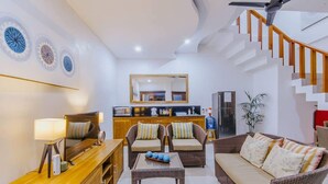 A modern living room with a sofa, chairs, a coffee table, and a TV. There is a kitchen area with a refrigerator and a microwave. A staircase with wooden railings is visible. - Secret Outlook Lembongan (Lembongan Island)