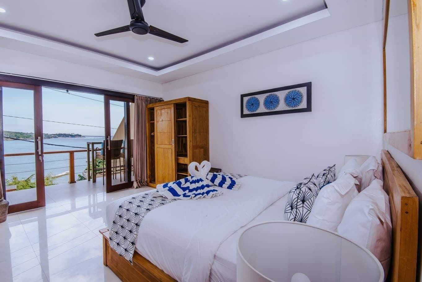 A bedroom with a large bed, a ceiling fan, a view of the sea, and a wooden wardrobe.